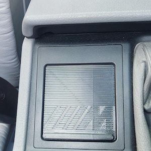 Peut inclure: Console intérieure de voiture avec une finition en grain de bois et une garniture en plastique gris. La console comporte un compartiment rectangulaire avec une surface texturée et les lettres "M" et "H" visibles. L'entourage est en cuir gris.
