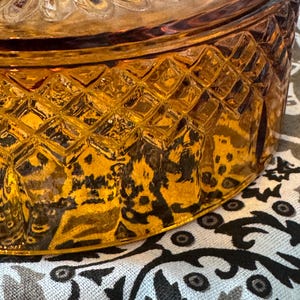May include: A round amber glass dish with a lid, featuring a textured diamond pattern around the side and a sunburst design on the top surface. The glass is translucent and reflects light, showing a clear geometric relief across its exterior.