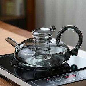 May include: A clear glass teapot with a rounded body, spout, and a curved handle. The teapot has a lid with a small knob and an internal infuser. It sits on a black electric hob.