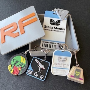 Könnte beinhalten: Eine Sammlung von Schlüsselanhängern und eine Box mit den Buchstaben "RF" in Orange. Schlüsselanhänger umfassen eine "Stella Montis Secure Access Keycard", eine Pistole und andere grafische Designs. Die Schlüsselanhänger sind silber und schwarz mit verschiedenen Farben.