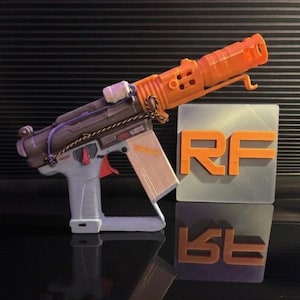 May include: A 3D-printed toy gun in orange, grey, and brown. The toy gun features a trigger, magazine, and barrel. The letters "RF" are displayed in orange on a silver square. The background is black.