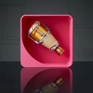 Könnte beinhalten: Ein gold- und silberfarbenes, zylindrisches Objekt mit beigefarbenem Mittelteil, das einem Musikinstrument oder einer Raketenkomponente ähnelt, wird in einem rosa quadratischen Behälter gezeigt. Das Objekt ist vor einem dunklen Hintergrund abgewinkelt.