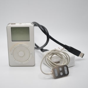 May include: A white digital music player with a rectangular screen and a circular click wheel interface. It is accompanied by a black charging cable, a white power adapter, and a white wired remote control with four small grey buttons.