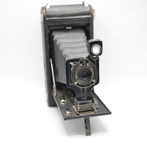 May include: A vintage black folding camera with a bellows mechanism, a circular lens assembly, and a small viewfinder. The front plate features the text 'No. 1A Autographic Kodak Jr.' and the lens housing includes the Kodak brand name and details.