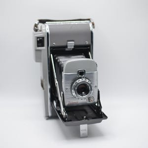 Puede incluir: Una cámara plegable Polaroid vintage con cuerpo gris y panel frontal plateado. La cámara cuenta con un fuelle negro, una lente circular con un anillo metálico y el nombre de la marca Polaroid impreso en rojo en la parte inferior delantera.