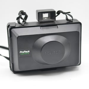 May include: A black Polaroid ProPack camera featuring a rectangular body with a central lens housing, a top-mounted viewfinder, and a black carrying strap. The front panel displays the text ProPack camera in white and green lettering.