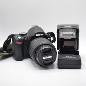 May include: A black Nikon digital SLR camera with a lens cap, a black camera strap with yellow Nikon branding, and a separate black Nikon Speedlight flash unit. The camera body features the Nikon logo on the front and the lens cap.
