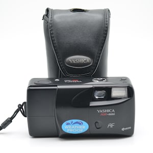 Peut inclure: Un appareil photo argentique 35 mm Yashica AW-mini noir avec flash intégré, objectif et un autocollant bleu Weather Proof sur le devant. L'appareil porte les inscriptions Yashica AW-mini et AF. Il est accompagné d'un étui de protection noir.