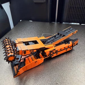 Op de afbeelding: Een oranje en zwart model van een mijnbouwmachine. Het model heeft een spijkerachtige roterende trommel aan één uiteinde en een transportbandachtige structuur aan de bovenkant. De machine staat op een zilveren oppervlak.