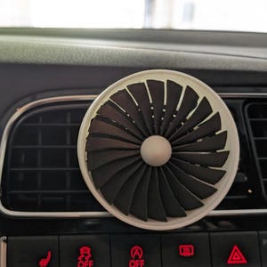 Könnte beinhalten: Ein Auto-Luftauslassdiffusor mit Turbinendesign. Der Diffusor ist schwarz und weiß, hat eine kreisförmige Form und eine zentrale Kugel. Er befindet sich auf einem schwarzen Armaturenbrett mit rot beleuchteten Knöpfen.