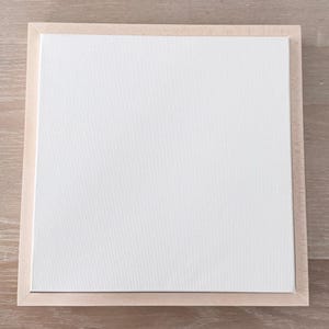 May include: A square blank white canvas mounted within a light-toned natural wood frame. The canvas surface features a visible woven texture, and the frame has a flat, smooth profile surrounding the entire perimeter of the white center panel.