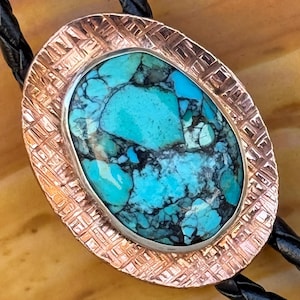 May include: A bolo tie featuring an oval-shaped centerpiece with a vibrant turquoise stone displaying dark matrix patterns. The stone is set within a textured copper-toned metal frame. The cord is made of black braided leather material.