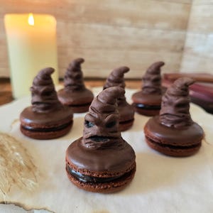 May include: Chocolate macarons shaped like the Sorting Hat from the Harry Potter series. The macarons are dark brown with a detailed hat design on top. They are arranged on a white, scalloped-edge plate, with a lit candle in the background.