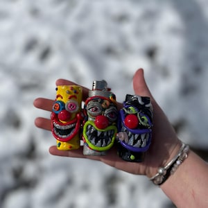 May include: Three custom-designed lighters with clown faces. The lighters are yellow, silver, and black, each with a unique, colorful clown face design. The clowns have red noses, exaggerated smiles, and various other facial features. The lighters are held in a hand against a snowy background.
