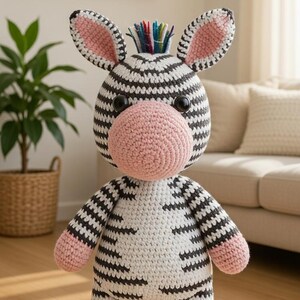 Peut inclure: Une peluche zèbre crochetée, rayée noir et blanc, avec des oreilles et un museau roses, et une crinière multicolore. Le jouet est posé sur un sol en bois. Jouet fait main.
