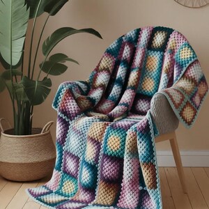 Può includere: Una colorata coperta all'uncinetto drappeggiata su una sedia. La coperta presenta un motivo geometrico nei toni del turchese, viola, giallo e rosa. I bordi sono rifiniti con un bordo azzurro. Una grande pianta in vaso e un cesto intrecciato sono sullo sfondo.