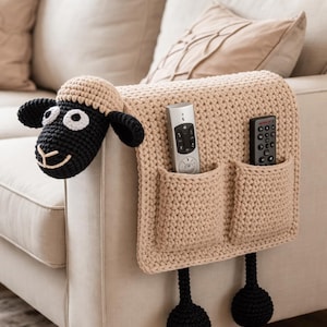 Könnte beinhalten: Ein beigefarbener, gehäkelter Schaf-Sofa-Arm-Organizer mit zwei Taschen, in denen sich zwei Fernbedienungen befinden. Das Schaf hat ein schwarzes Gesicht, Ohren und Beine. Der Organizer ist am Arm eines beigefarbenen Sofas befestigt.