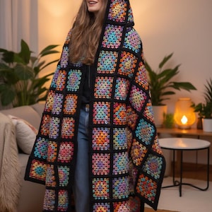 Peut inclure: Une couverture longue à capuche en crochet, motif granny square. La couverture présente une bordure noire avec des carrés colorés dans les tons de rose, orange, jaune, bleu et violet. La couverture est portée sur les épaules.