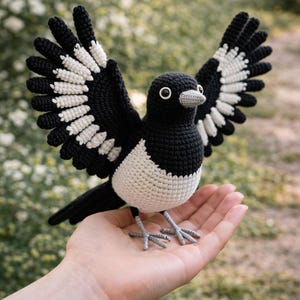 May include: A handmade crochet magpie bird with outstretched wings, showcasing a detailed black and white pattern. The bird's body is white, with black wings and tail. The bird is perched on a hand, with a blurred green background.