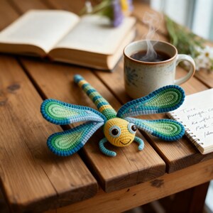 Puede incluir: Un juguete de libélula hecho a mano con ganchillo en amarillo, azul y verde, sobre una mesa de madera. La libélula tiene alas grandes y detalladas y una cara sonriente. Una taza de café, un libro abierto y un bloc de notas también están presentes.