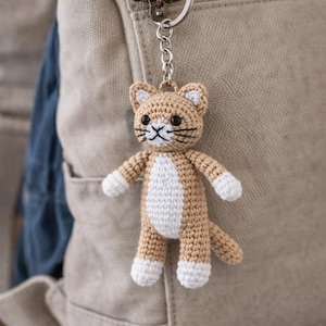 Peut inclure: Un porte-clés chat crocheté, de couleur beige et blanc, suspendu à un porte-clés argenté. Le chat a des moustaches noires et des pattes blanches. Le porte-clés est accroché à un sac à dos beige.