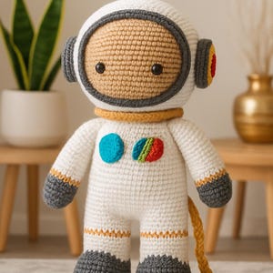 Peut inclure: Poupée d'astronaute au crochet faite à la main. La poupée est principalement blanche avec des accents gris et jaunes. Elle présente un casque détaillé, un écusson bleu et arc-en-ciel et un cordon jaune. La poupée mesure environ 25 cm de haut.