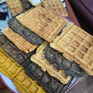May include: A stack of rectangular waffles in various colours including golden yellow, dark charcoal grey, and light beige. The waffles feature a classic grid pattern with deep indentations and a crisp, textured surface, arranged in a pile on a plate.