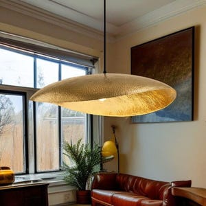 Può includere: Un grande lampadario a sospensione in metallo martellato color oro. La luce ha una forma ovale ampia e poco profonda e pende da un cavo nero. L'interno della luce è di un colore oro brillante. L'apparecchio si trova in una stanza con una finestra e un divano in pelle.