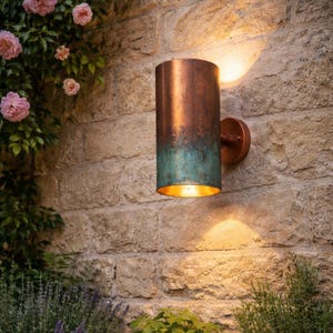 May include: A cylindrical outdoor wall sconce with a copper finish and a turquoise patina at the bottom. The sconce is mounted on a stone wall and emits a warm light. The fixture is approximately 12 inches tall.