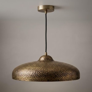 May include: A pendant light featuring a wide, dome-shaped shade with a hammered, antique brass finish. The shade is suspended by a thin black cord attached to a matching circular brass ceiling canopy, creating a clean and industrial aesthetic.