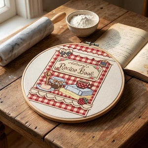 May include: A marble rolling pin with wooden handles, a bowl of flour, and an open vintage cookbook rest on a rustic wooden table. A cross-stitch hoop features a recipe book design with the text "Sidney's Recipe Book".