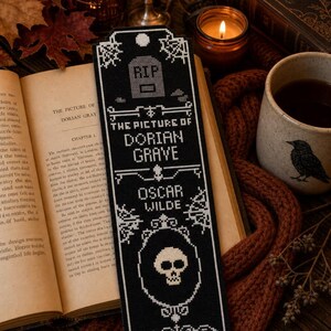 May include: A black and white cross-stitch bookmark featuring a tombstone with the text RIP, the title THE PICTURE OF DORIAN GRAVE, and the author OSCAR WILDE, finished with a skull graphic and spiderweb details at the top and bottom corners.