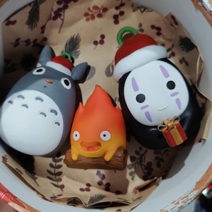 Könnte beinhalten: Drei kleine Charakterfiguren in einer runden Schachtel. Eine ist ein grauer Totoro mit Weihnachtsmütze, eine ist ein leuchtend orangefarbener Calcifer auf einem Holzscheit und eine ist ein schwarz-weißer Ohngesicht-Charakter mit Weihnachtsmütze.