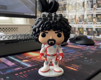 Può includere: Una statuetta in vinile Funko Pop di un personaggio con capelli neri ricci, pizzetto e tuta spaziale bianca e rossa. La statuetta è su una base bianca. Lo sfondo include un monitor del computer, tastiera, mouse e un muro di altre statuette Funko Pop.