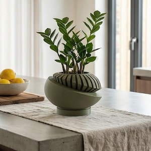 May include: A modern, olive green ceramic planter with a unique, geometric design, holding a lush green plant. The planter sits on a countertop with a neutral-toned runner, alongside a bowl of lemons and a wooden cutting board. The kitchen setting is bright and airy.