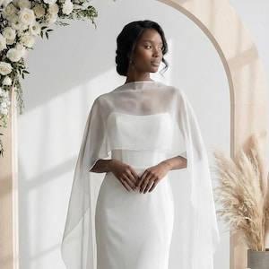 Op de afbeelding: Een witte trouwjurk met een doorschijnende cape. De jurk heeft een nauwsluitend lijfje en een zwierige rok. De cape bedekt de schouders. De achtergrond omvat een houten boog en bloemstukken. De stijl is elegant.