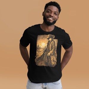 Puede incluir: Camiseta negra con un gráfico en tonos sepia de un vaquero en un paisaje desértico, con cactus y un sol poniente. El vaquero lleva sombrero y abrigo largo, sosteniendo un arma. El diseño tiene un estilo vintage y artístico.