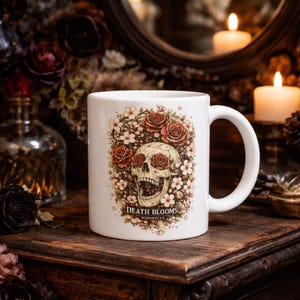 Puede incluir: Taza de cerámica blanca con un diseño de calavera, rosas en las cuencas de los ojos y detalles florales. La taza tiene el texto "DEATH BLOOMS" y "NIPONELLA". La taza está sobre una superficie de madera.