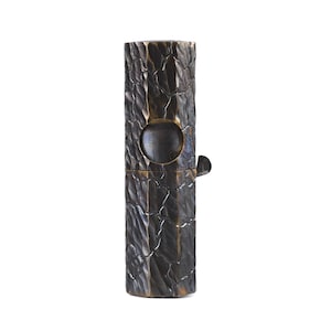 Pode incluir: Um recipiente cilíndrico castanho escuro com um exterior texturizado semelhante a casca de árvore. Possui uma abertura circular e uma pequena alça articulada. O recipiente parece ser feito de metal e tem uma estética rústica e artesanal.