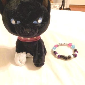 May include: A black plush cat with blue eyes and a red collar with white polka dots. A beaded bracelet with the word "SC0URGE" is next to the cat. The bracelet has pink, blue, white, black, and purple beads.