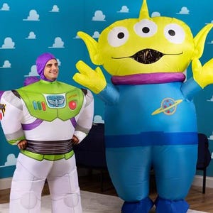 Puede incluir: Dos disfraces inflables, uno que se asemeja a Buzz Lightyear con detalles verdes y blancos, y el otro un extraterrestre con tres ojos y un cuerpo azul. El fondo es una pared azul con formas de nubes blancas.