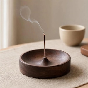 以下が含まれることがあります： 中央に円錐形のデザインが施された、濃い茶色の木製お香立て。 燃えているお香スティックから煙が立ち上っています。 背景にはベージュのセラミックボウルがあります。 ホルダーはベージュの表面にあります。
