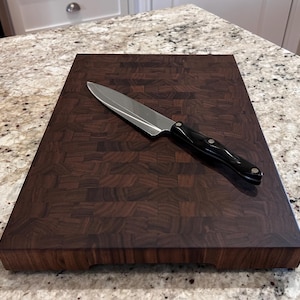 May include: A rectangular end-grain wooden chopping board with a dark brown, multi-block pattern. A stainless steel chef's knife with a black handle and three silver rivets rests diagonally across the centre of the board's surface.