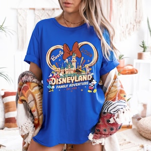 May include: A royal blue t-shirt featuring a Disneyland graphic. The design includes a castle, Mickey and Minnie Mouse, and the words "Bestie 2020" and "Family Adventure Est. 1955". The shirt has rolled-up sleeves.