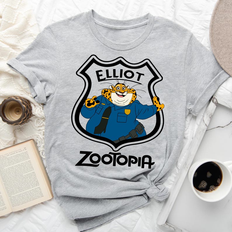 May include: A light grey t-shirt with a cartoon leopard character in a police uniform. The character is inside a badge that says "ELLIOT" at the top and "ZOOTOPIA" at the bottom in black text.