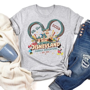 May include: A heather gray t-shirt featuring a Disneyland graphic. The design includes Mickey and Minnie Mouse, Donald Duck, and other characters, along with the text "Disneyland Family Adventure + Est. 1955" and the year "2020".