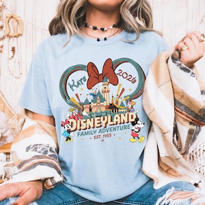 May include: Light blue t-shirt featuring a Disneyland graphic. The design includes a castle, Mickey Mouse, Minnie Mouse, Donald Duck, and other characters. The text reads "Kaye 2026" and "Disneyland Family Adventure Est. 1955".