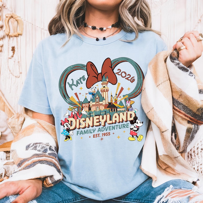 May include: Light blue t-shirt featuring a Disneyland graphic. The design includes a castle, Mickey Mouse, Minnie Mouse, Donald Duck, and other characters. The text reads "Kaye 2026" and "Disneyland Family Adventure Est. 1955".