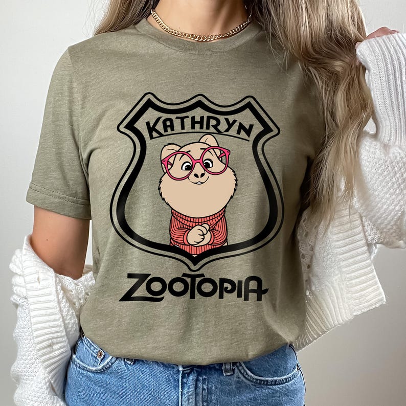 May include: An olive green t-shirt with a cartoon character from Zootopia wearing glasses and a sweater, inside a shield with the name "Kathryn" at the top. The word "Zootopia" is printed below the shield in a stylized font.