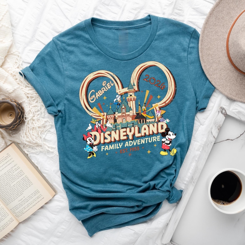 May include: Teal t-shirt with a Disneyland graphic featuring Mickey and Minnie Mouse, a castle, and the text "Gabriel 2020 Family Adventure Est. 1955". The design is in a Mickey Mouse ear shape.